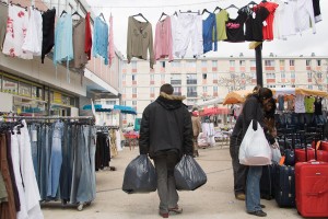 un homme de dos marche au milieu d'un marché au vêtements , en tenant deux sacs, devant un immeuble d'habitation.
