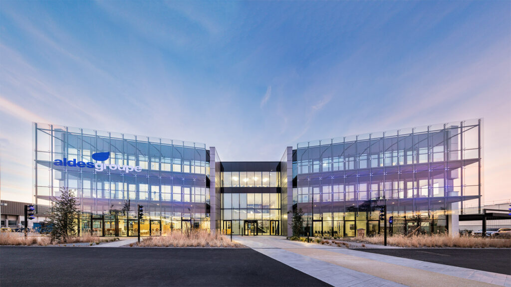 Image d'un grand immeuble de bureaux en verre avec un ciel bleu en arrière-plan. Le bâtiment est le siège social du groupe Aldes, une entreprise française spécialisée dans la ventilation et la climatisation. Le logo Aldes est visible au sommet du bâtiment.