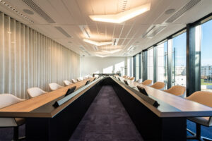 Image d'une salle de réunion du siège d'ALDES avec une longue table en bois et des chaises confortables. La salle est éclairée par de grandes fenêtres qui donnent sur une ville.